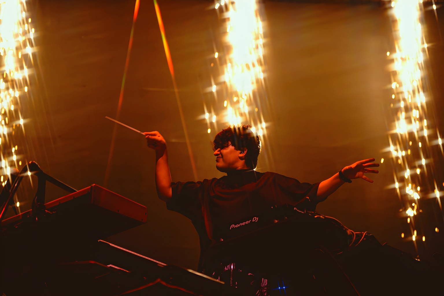Shashwat Sachdev conducts with a baton and both arms extended beneath golden stage sparks.